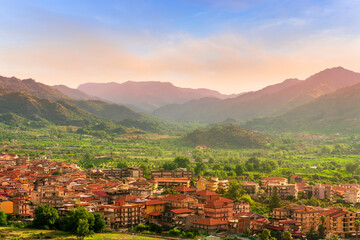 Obraz premium wonderful landscape of sunset valley town with beautiful fields, gardens, green plantations among great mountains and scenic evening cloudy sky on background of landscape