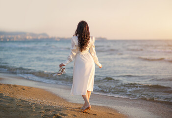 A girl on vacation walks on a sandy beach by the sea in a white dress.