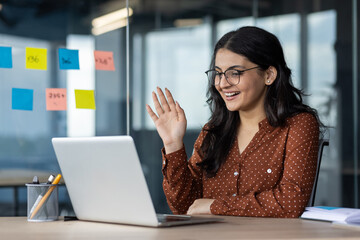 Latina businesswoman in office enjoying video call using laptop, smiling and waving. Highlights remote work, technology, professional connection. Modern office setting with creative notes on wall.