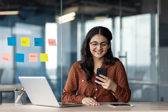 Latino businesswoman in office, multitasking with smartphone and laptop. Smiling, showcasing technology use and effective communication. Bright workspace with sticky notes adds creative vibe.