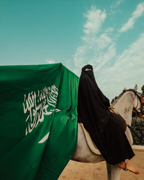 A woman in a burqa, covering her face, sits on a horse in front of the Saudi Arabian flag