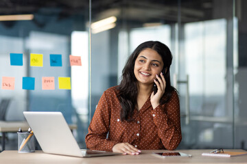 Latino businesswoman smiling during phone call at office desk. She's using a smartphone while...