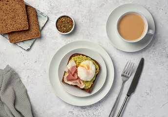 Food photography of nutritional breakfast, sourdough rye bread with mashed avocado, prosciutto, bacon, boiled poached egg, tea with milk