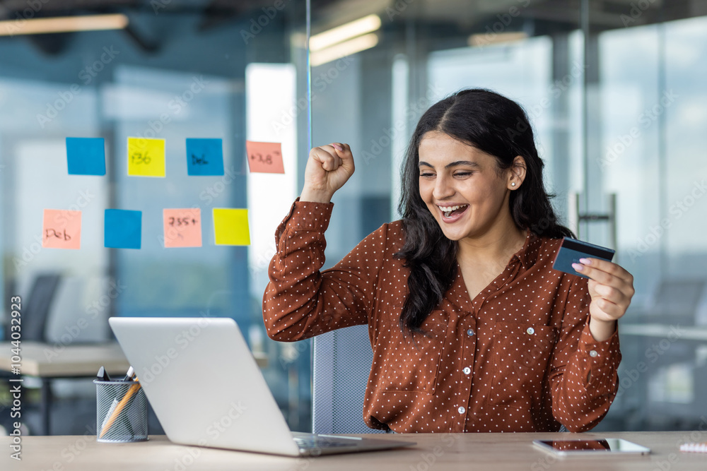 Poster Latino businesswoman excited during online transaction with credit card using laptop in office. Empowerment, success, happiness captured. Phone visible on desk, indicating multitasking.