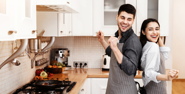 Having Fun. Carefree beautiful european couple wearing aprons dancing in the kitchen, cooking delicious dinner