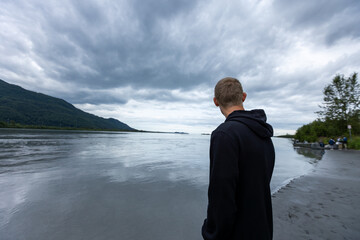 Fototapeta premium An explorer stands quietly on the bank of Alaska’s Knik River, gazing at the serene, icy waters under the soft glow of a cold, crisp morning.