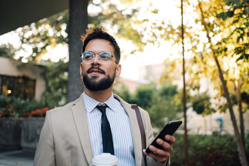 Confident businessman holding coffee walking using smartphone outdoors