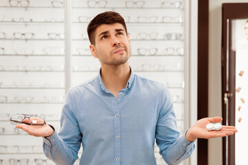 Pensive man choosing between eyeglasses and contact lenses standing in the optical shop