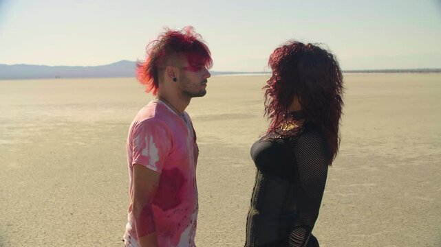 Redhead man in blood-stained shirt, and gothic woman approach each other on a sunlit, desolate desert plain and almost share a kiss. Punk guy and Goth girl about to kiss in the middle of a vast desert