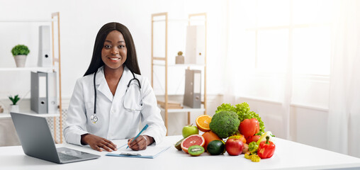 Smiling black woman dietologist working with laptop in modern office, having fresh fruits and vegetables on table, copy space