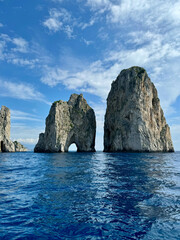 Landscape of the Faraglioni rocks surrounded by the blue water of the Mediterranean Sea with clouds in the sky