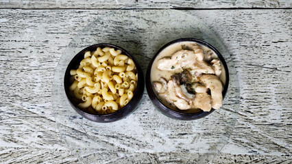 Two Ceramic Bowls with Macaroni Pasta and Creamy Mushroom Chicken on Glass Plate with Rustic Wooden Background