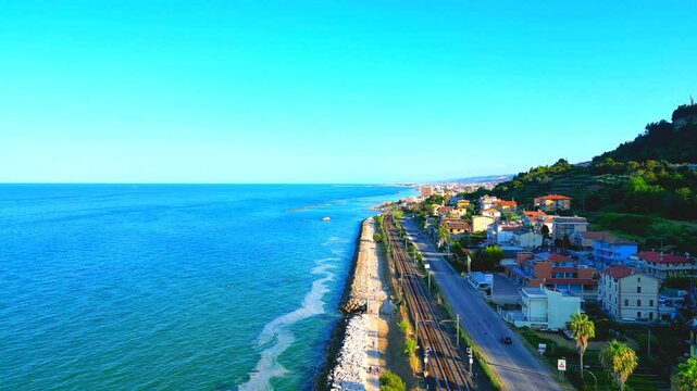 Soaring aerial push-in shot from Cupra Marittima, with vast finely rippling blue Adriatic Sea skimming the stony seawall, breakwaters, rocks, swaths of promenade, railway and main road, cars, edifices