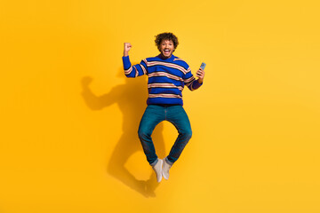 Full length photo of handsome young guy jump hold device winning dressed stylish blue striped garment isolated on yellow color background