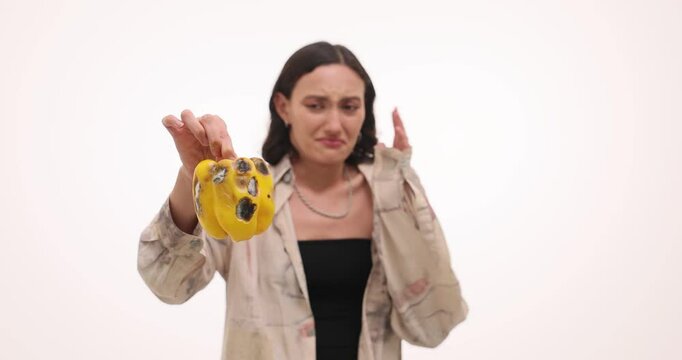 Young woman holds rotten vegetable expressing disgust. Lady turns away smelling spoiled bell pepper with strong reaction of revulsion slow motion