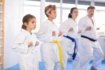 Fototapeta premium Focused preteen girl in kimono standing in attacking stance, practicing punches and kicks as part of kata along with brother, mother and father. Concept of family joint sports activity..