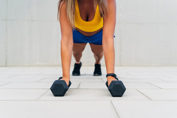 Sportswoman doing push-ups holding dumbbells in a white urban background
