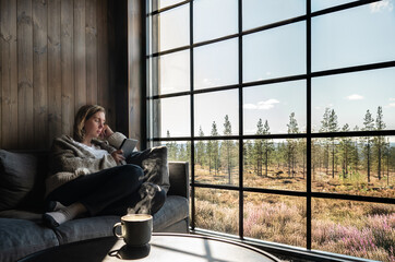 A woman sitting comfortable reading on a couch by a large glass window in a cabin with a hot drink beside her, absorbed in an e-reader book while enjoying the tranquil view of a forest outside