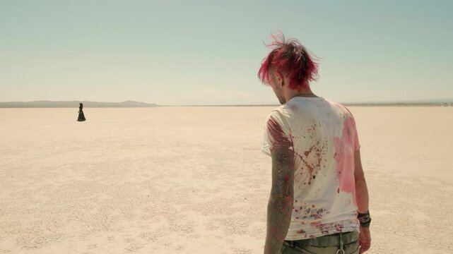 Young man with red dyed hair and blood-covered t-shirt slowly walks through vast desert, approaching a lonely female figure in black. Young Punk guy approaches Goth girl in a desert.