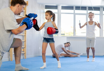 Focused cute preteen girl wearing boxing gloves working out with father during self-defense course,...