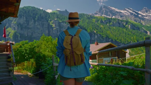An attractive girl in a blue shirt and a fashionable hat spends her holidays alone, traveling through the Alpine meadows. Tourist with a backpack travels through Switzerland with incredible views
