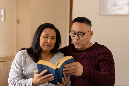 Multiracial senior couple reading book together on cozy couch at home, enjoying time