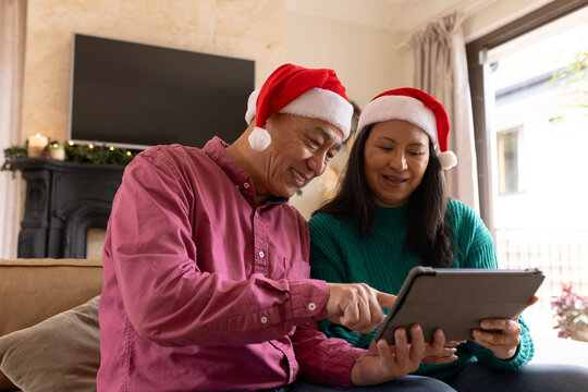On video call, multiracial senior couple wearing santa hats using tablet at home, enjoying christmas