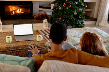 multiracial couple relaxing by fireplace, watching laptop near Christmas tree at home, copy space