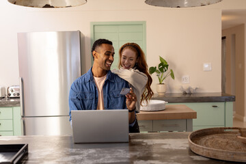 multiracial couple shopping online for Christmas gifts in cozy kitchen, smiling happily, at home