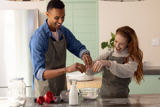 Multiracial couple baking together in kitchen, cracking eggs and smiling joyfully, at home - Powered by Adobe