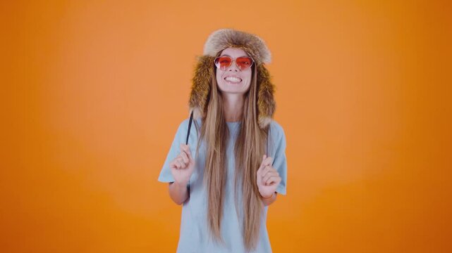 A girl wearing funny pink glasses and a ushanka hat smiles at the camera, showcasing her playful side. The bright orange background adds a lively touch to her cheerful expression