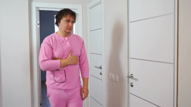 Sleepy, exhausted man in pink pajamas, disheveled, languidly leaves bedroom with eyes closed, scratching his chest with his hand. No energy, motivation, vitality Morning lethargy, difficulty waking up