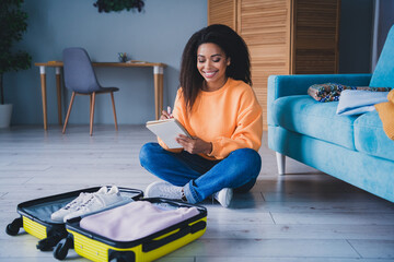 Full length photo of lovely young girl sit floor pack suitcase write list dressed orange garment...