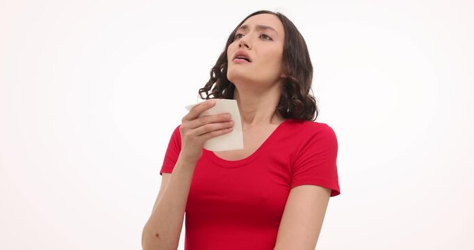 Ill woman blows nose with tissue suffering from cold symptoms on white background. Brunette female suffers from virus feeling discomfort in nose