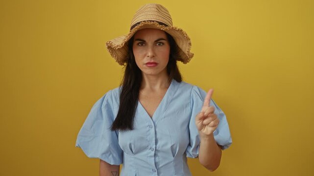 Young woman in a light blue dress and straw hat gesturing no with her finger against an isolated yellow background