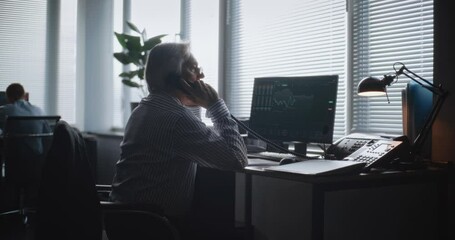 In modern office: Senior financial specialist talks to business client using landline phone, analyzes paper documents. PC screen shows equity market and shares. International stock exchange company. - Powered by Adobe