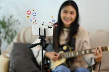 Young woman playing guitar during live streaming on smartphone at home