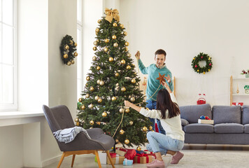 Happy joyful smiling young couple in love decorating Christmas fir tree together standing in festive living room at home preparing for New Year celebration. Winter holidays concept.