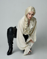 full length portrait of beautiful female model, wearing vintage gothic fantasy ruffle blouse white shirt, leather pants, blonde hair holding knife weapon. sitting pose, isolated on studio background.