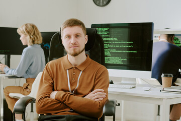 Medium shot of young Caucasian programmer sitting with arms crossed at workplace in IT office...
