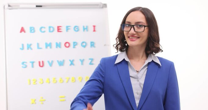 Female teacher stands by Latin alphabet chart giving thumbs up on white background. Tutor radiates positivity reflecting support in learning