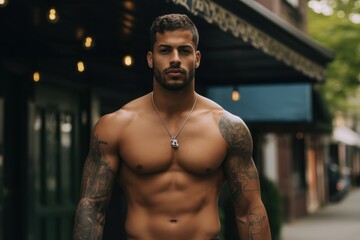 A man with a necklace and tattoos on his arms stands in front of a building