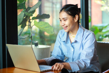 excited asian woman looking at laptop on wooden table talking on video chat, online meeting, sitting in cafe near window with green plant