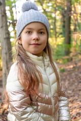 A little girl of 6 wearing a grey hat and jacket smiles in a sunlit autumn forest.