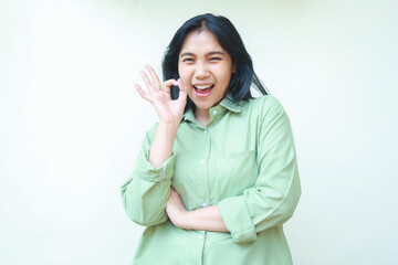 delighted asian woman with dark hair wearing green over size clothes showing ok signs with folding arms looking at camera screaming satisfied isolated on white background