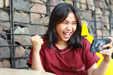 carefree asian woman celebrating winning playing online game competition using smartphone with raising fist say yes gesture sitting in outdoor cafe