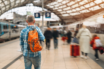 A traveler stands with an orange backpack at a bustling train station during a sunny afternoon