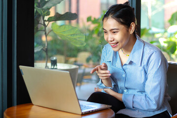 excited asian woman sitting in cafe talking on laptop have video chat, online meeting, with pointing finger wearing striped blue shirt
