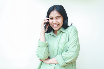 carefree asian woman with dark hair wearing green over size shirt enjoy talking on smartphone with arms folded, playful stylish female looking at camera, standing over isolated white background