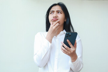 curious asian young business woman looking up wondering and thinking while using mobile phone app with hand on chind, surfing internet, wear white formal suit standing over white background